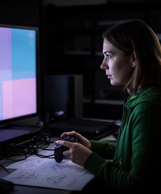 Woman game reviewer comparing console performance notes beside a television in a dark editorial room